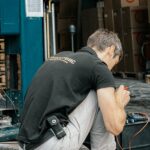 Technician crouching while repairing machinery at an outdoor warehouse with boxes in background.
